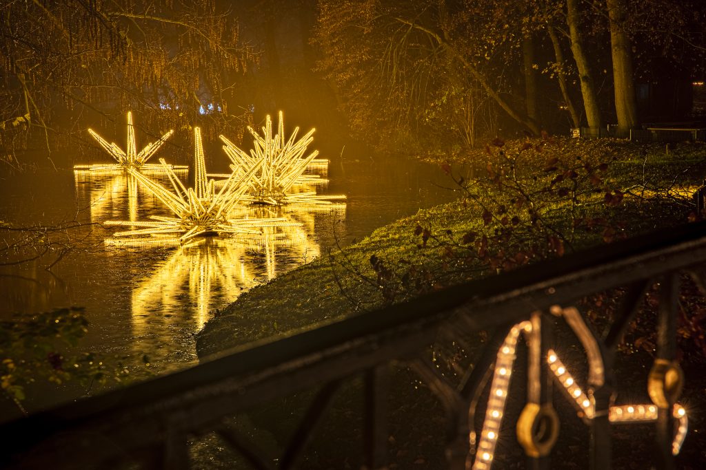 Große sternförmige Lichtinstallationen schweben nachts auf einem Teich. Im Vordergrund sind Bäume und ein geschmücktes Geländer zu sehen. Große sternförmige Lichtinstallationen schweben nachts auf einem Teich. Im Vordergrund sind Bäume und ein geschmücktes Geländer zu sehen.