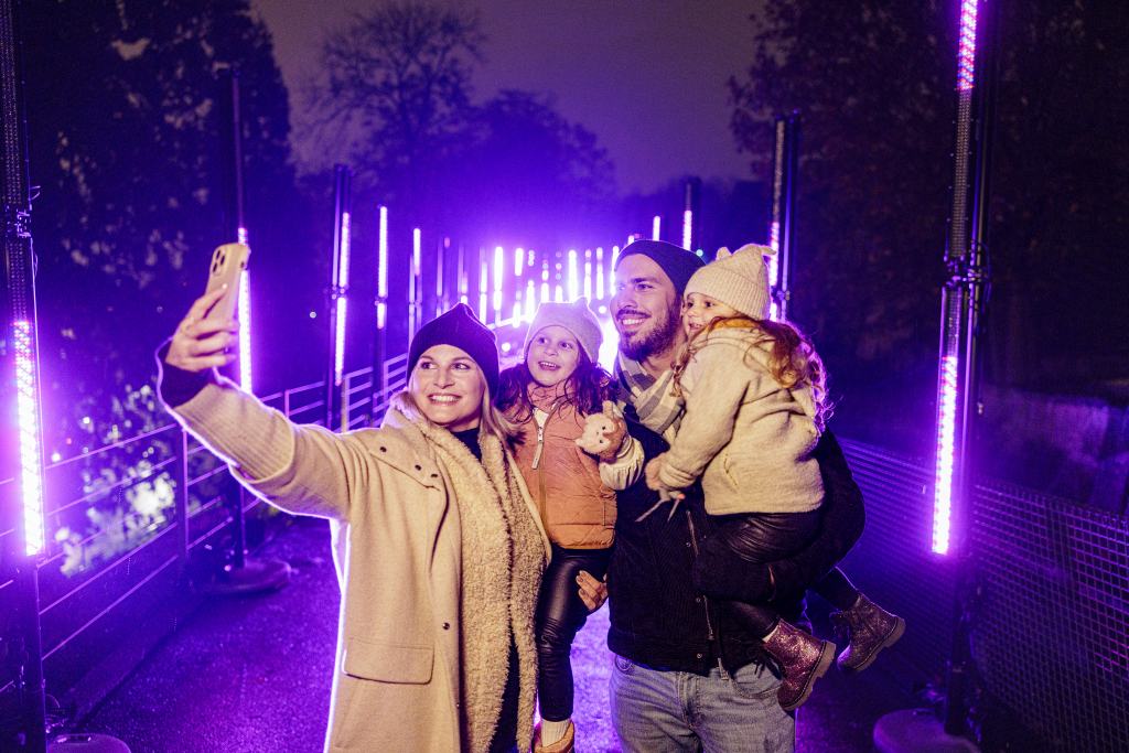 Eine vierköpfige Familie macht auf einer Brücke bei Nacht ein Selfie. Sie sind von hellen, lila Lichtern umgeben und tragen warme Winterkleidung.