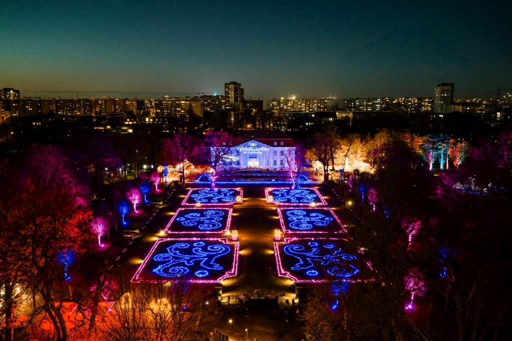 Ein formaler Garten ist nachts bunt beleuchtet. Im Hintergrund stehen ein Herrenhaus und dahinter hohe Gebäude der Stadt.
