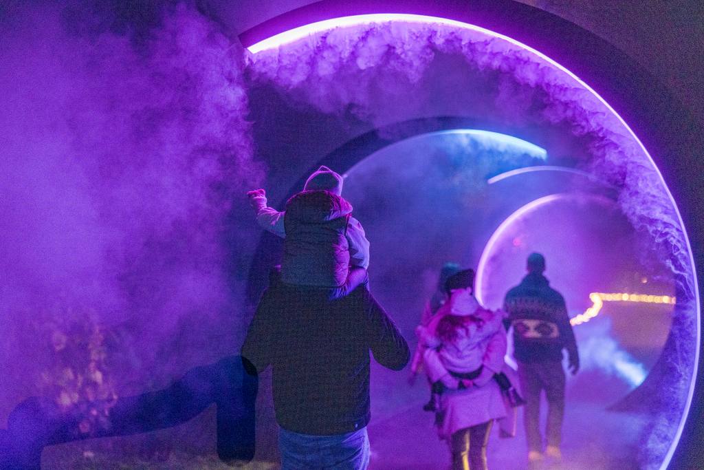Menschen gehen durch einen nebligen Tunnel mit violetten Lichtern. Eine Person trägt ein Kind auf den Schultern.